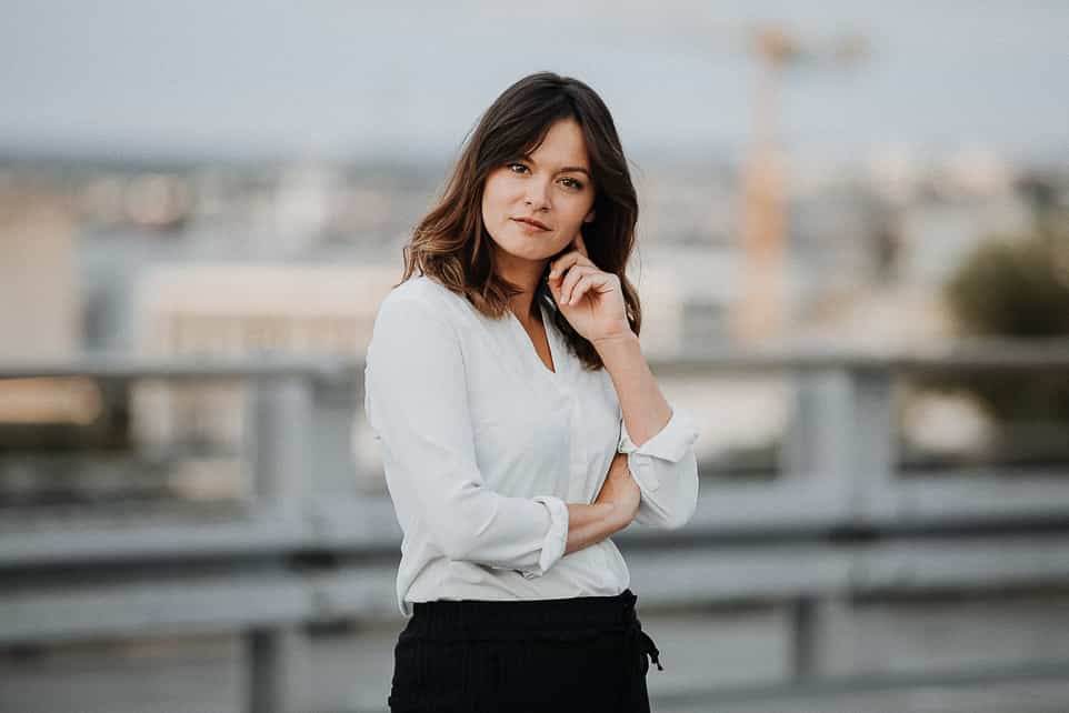 Modernes Bewerbungsfoto einer charismatischen jungen Frau mit weißer Bluse und verschwommener Skyline im Hintergrund