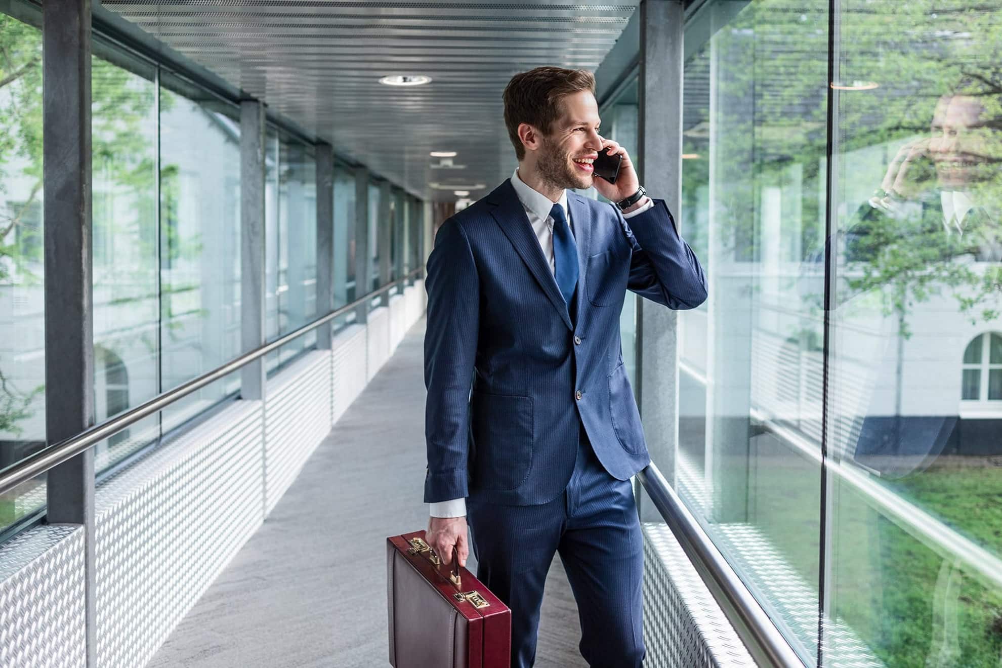 Moderner, lachender junger Business Mann im blauen Anzug der einen Flur mit Glaswänden entlang läuft während der telefoniert und einen Koffer trägt.