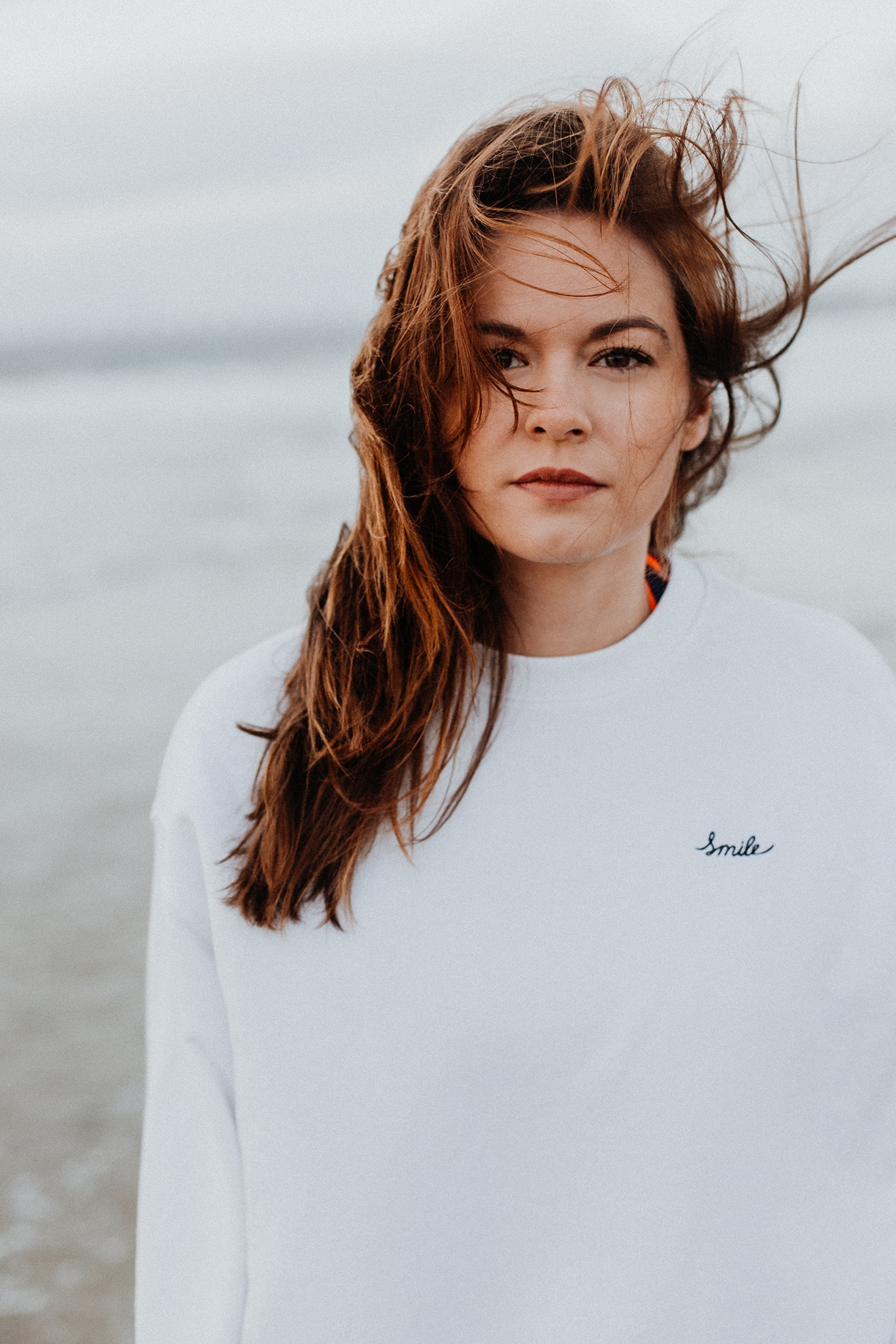 Portrait von Diane Willems im weißen Pullover am windigen Meer 