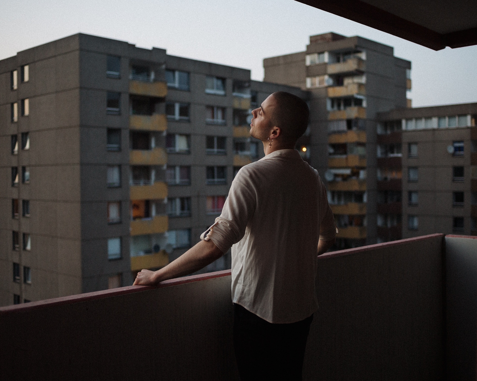 Junger Mann blickt von einem Balkon in dunkler Lichtstimmung umgeben von Plattenbauten