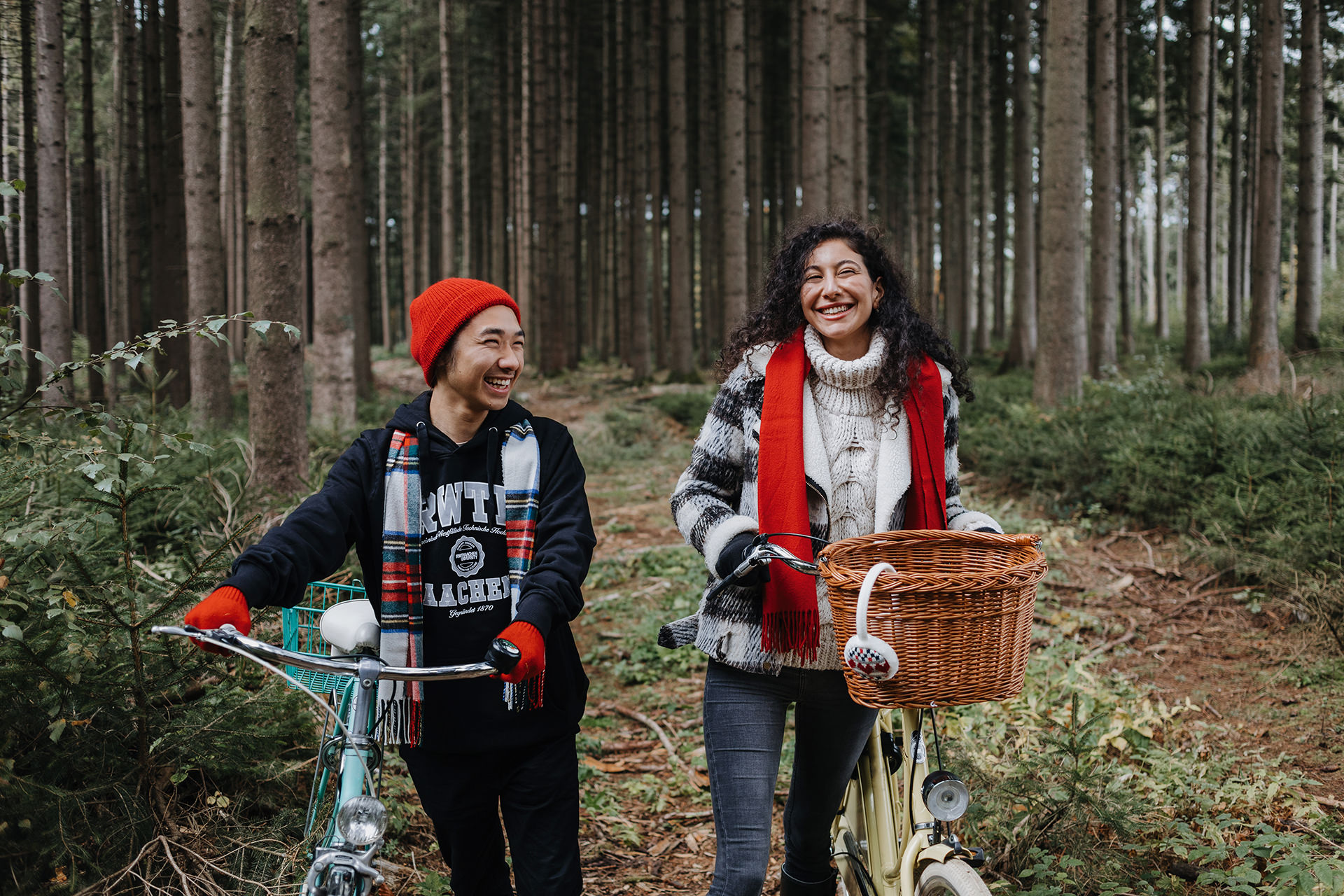 Junges lachendes Paar mit rotem Schal und Mütze schiebt die Fahrräder im Winter durch den Wald