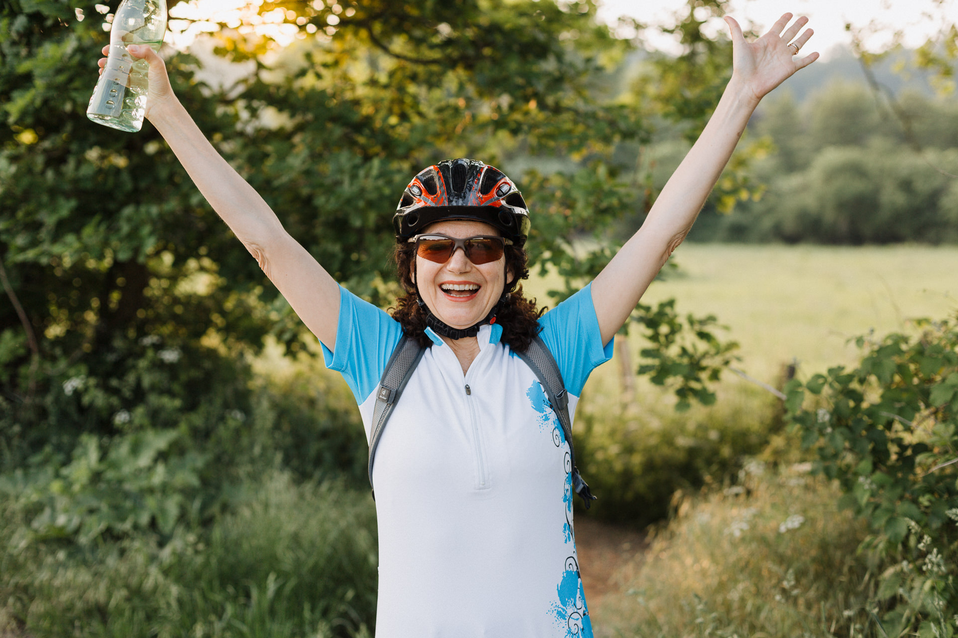 Glückliche Radfahrerin mit Helm in der Natur wirft die Arme freudig in die Luft.