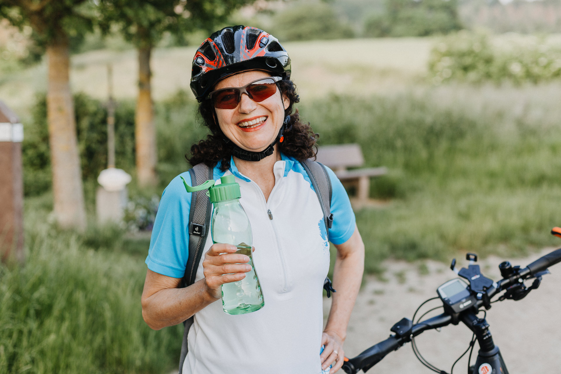 Sympathische Radfahrerin mit Fahrrad im Hintergrund mittleren Alters hält eine Trinkflasche in der Hand.
