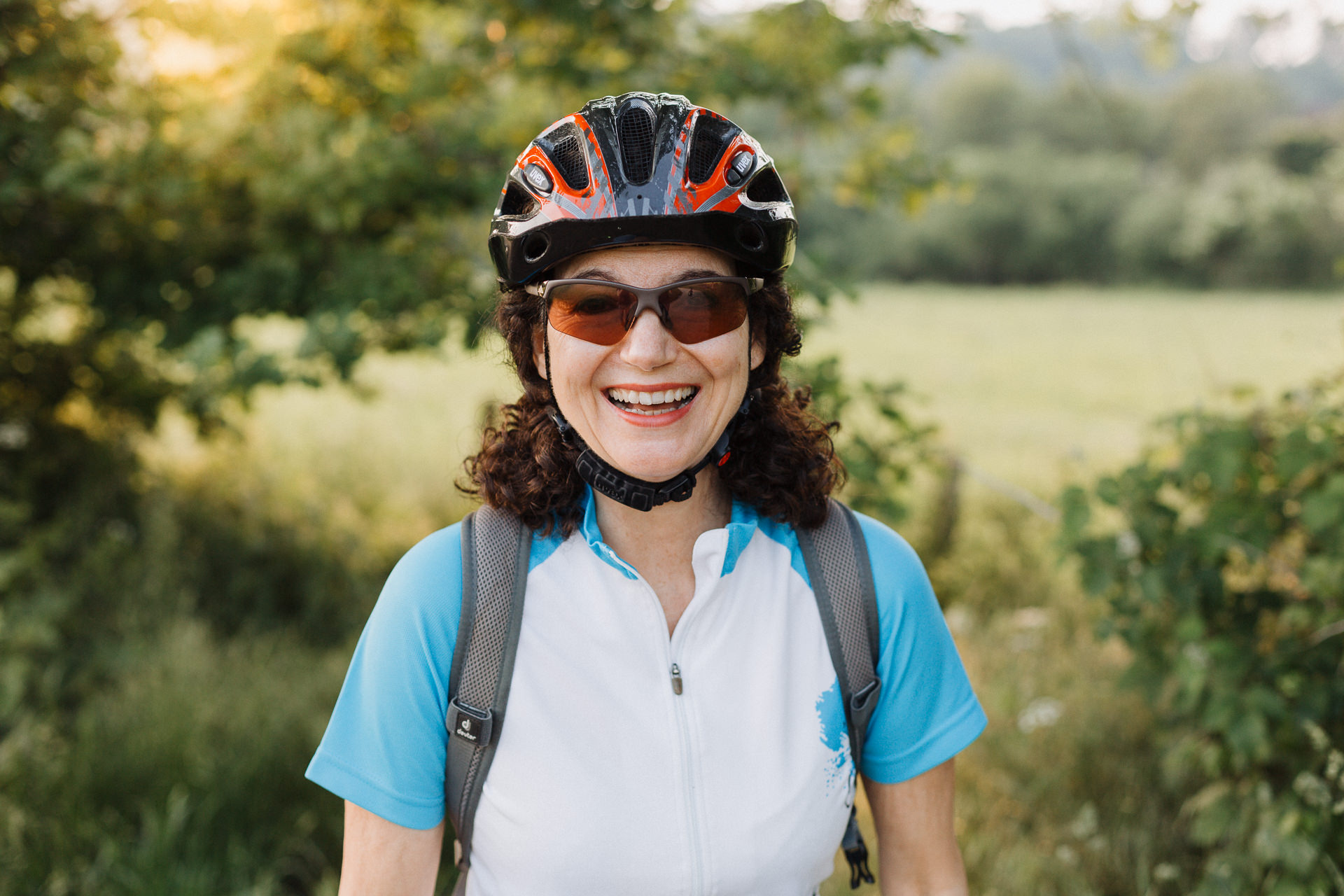 Lachende sympathische Radfahrerin mit Helm mittleren Alters in grüner Natur und Sonnenuntergang im Hintergrund